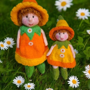 May include: Two handmade knitted dolls in a grassy setting with daisies. The dolls wear orange and yellow dresses with green accents, matching hats, and green shoes. The dolls have rosy cheeks and are decorated with daisy details.