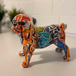 Peut inclure: Une figurine en céramique colorée de style graffiti représentant un carlin. La figurine est peinte dans des tons d'orange, de bleu, de jaune et de noir, avec une variété de motifs et de designs abstraits.
