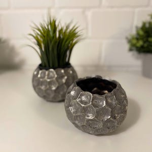 Può includere: Due vasi in ceramica color argento con un design testurizzato a forma di nido d'ape. I vasi sono rotondi e hanno una superficie leggermente irregolare.