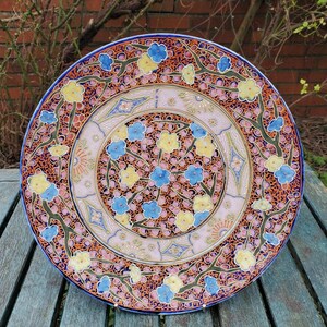 Può includere: Un grande piatto rotondo in ceramica con un design floreale blu, rosa, giallo e arancio. Il piatto ha un bordo rialzato e un motivo dettagliato di fiori e foglie.