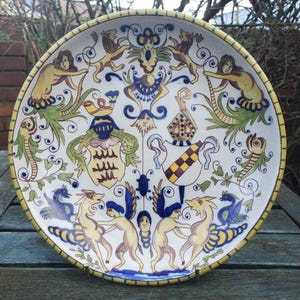Könnte beinhalten: Ein dekorativer Keramikteller mit weißem Hintergrund und gelbem Rand. Der Teller zeigt ein detailliertes, farbenfrohes Design mit Fabelwesen, heraldischen Symbolen und floralen Elementen. Der Durchmesser des Tellers beträgt ca. 30 cm.