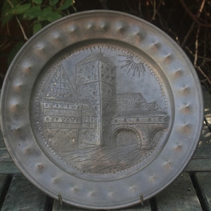 Peut inclure: Une assiette en étain avec un motif en relief d'une ville médiévale avec une tour et un pont. L'assiette a un bord surélevé avec un bord perlé.