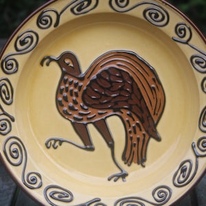 Op de afbeelding: Een keramisch bord met een bruin en geel ontwerp. Het bord heeft een gestileerde vogel met bruine en zwarte veren. De rand van het bord is versierd met een bruin swirl-patroon.