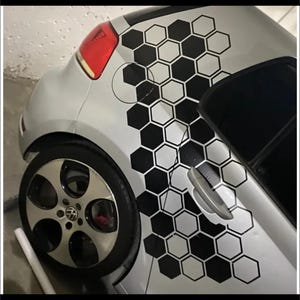 Puede incluir: Un coche blanco con calcomanías de panal de abeja negras en el lateral. Las calcomanías son un patrón repetitivo de hexágonos. El coche tiene una rueda negra con una llanta plateada. El texto "KITS" está impreso en negro en la parte inferior de la imagen.
