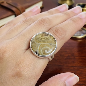 Pode incluir: Um anel com uma grande pedra de coral fossilizado circular e polida, com intrincados padrões de redemoinho em tons bege e castanho-claro, engastada numa banda de metal prateado liso com um design de haste dividida.