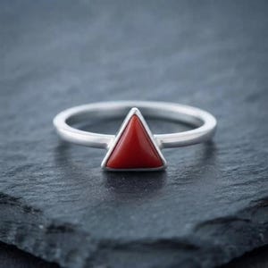 Peut inclure: Une bague en argent avec une pierre rouge lisse de forme triangulaire sertie au centre. L'anneau est en argent poli avec un profil arrondi, et la pierre est d'une couleur rouge opaque et unie avec une surface plate et des bords biseautés.