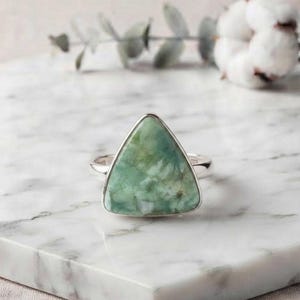 Peut inclure: Une bague ornée d'une grande pierre triangulaire verte aux veines marbrées blanches, montée sur un anneau en argent poli. La pierre présente une surface lisse et plate avec des variations de couleurs naturelles allant du vert sauge clair au foncé.