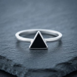 May include: A silver ring featuring a central triangular focal point. The triangle is filled with a smooth, solid black inlay. The band of the ring is polished silver with a rounded profile, creating a clean and geometric design for the finger.