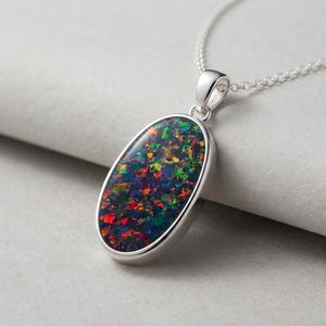 May include: An oval-shaped pendant necklace featuring a dark base stone with vibrant, multi-colored flecks of red, green, and blue. The stone is set in a polished silver frame and hangs from a delicate silver chain. The metal bail is marked 925.