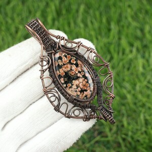 Op de afbeelding: Handgemaakte hanger met een gespikkelde edelsteen gezet in een koperdraadframe. De steen heeft een donkergroene basis met bruine en bruinachtige vlekken. Het draadwerk is ingewikkeld geweven, waardoor een decoratieve, bladvormige vorm ontstaat.