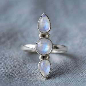 May include: A silver ring featuring three moonstones arranged vertically. The center stone is round, while the top and bottom stones are pear-shaped. Each stone displays a translucent, iridescent blue glow set within a polished silver metal band.