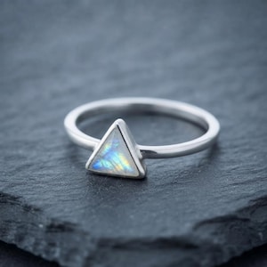 Peut inclure: Une bague en argent ornée d'une pierre de lune triangulaire sertie dans une monture en métal lisse. La pierre précieuse présente des reflets irisés bleus et jaunes sur un anneau en argent poli formant une boucle circulaire complète.