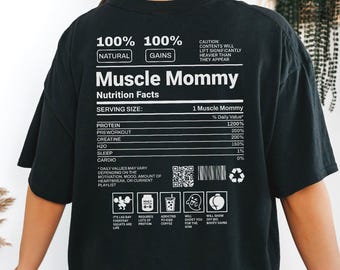 Puede incluir: Camiseta negra de gran tamaño con el texto "Muscle Mommy" y un diseño de etiqueta de información nutricional. La etiqueta incluye detalles humorísticos sobre proteínas, pre-entrenamiento, creatina y sueño. La camiseta también tiene el texto "100% Natural Gains".