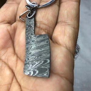 Könnte beinhalten: Ein kleiner, gemusterter Metall-Schlüsselanhänger in Form eines Hackmessers mit einem silbernen Schlüsselring. Das Metall hat ein wirbelndes, marmoriertes Aussehen in Grautönen. Der Schlüsselanhänger wird in einer Hand gehalten, was auf seine Größe und Tragbarkeit hindeutet.