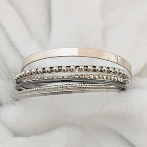 May include: A collection of silver bracelets with diverse styles. One is a plain band, others have small beads, textured detailing, and a black leather-look band. The bracelets are presented on a soft, white, textured background.