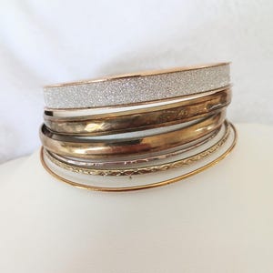 May include: A collection of metal bangles in various colours. The top bangle is rose gold with a silver glitter band. Below are gold, silver, and bronze bangles in different widths and textures. The bangles are stacked on a white surface.