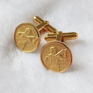 May include: A pair of gold cufflinks. Each cufflink has a circular design with a relief of a person in a dynamic pose. The cufflinks have a classic, elegant design, suitable for formal wear. The background is a soft, white fabric.
