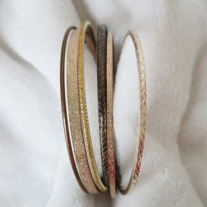 May include: A selection of six assorted bangles in various metallic finishes. The bangles include shades of gold, rose gold, bronze, and silver. Some have textured surfaces, while others are smooth. The bangles are stacked together.