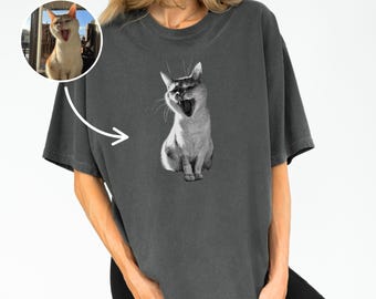 May include: Dark gray t-shirt featuring a black and white image of a yawning cat. A smaller, color image of the cat is in a circle in the upper left corner. The cat is sitting with its mouth wide open.