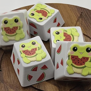Könnte beinhalten: Eine Sammlung weißer, quadratischer Tastenkappen mit einem Cartoon-Frosch-Design. Die Frösche sind hellgrün und halten Wassermelonenscheiben. Rote Wassermelonenscheiben sind ebenfalls auf den Seiten der Tastenkappen abgebildet.