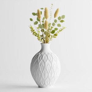 May include: A white ceramic vase with a textured leaf pattern, holding a bouquet of dried yellow and green foliage. The vase has a rounded shape and sits against a white background, creating a minimalist aesthetic.