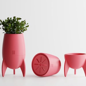 Può includere: Un set di tre fioriere a forma di razzo color corallo. Una fioriera contiene fogliame verde. Le altre due fioriere sono vuote, una è sdraiata su un lato. Le fioriere hanno tre gambe e un design dettagliato.