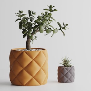 Könnte beinhalten: Zwei dekorative Blumentöpfe, einer groß und einer klein, jeweils mit einem Steppmuster. Der größere Topf ist senfgelb und enthält eine Jade-Pflanze. Der kleinere Topf ist grau und enthält eine Sukkulente.