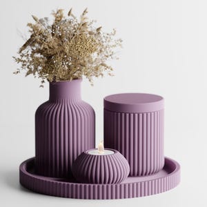 Op de afbeelding: Een decoratieve set in een gedempte paarse tint, met een geribbelde vaas met gedroogde bloemen, een bak met deksel, een kleine kandelaar met een brandende kaars en een rond dienblad. Op een witte achtergrond.