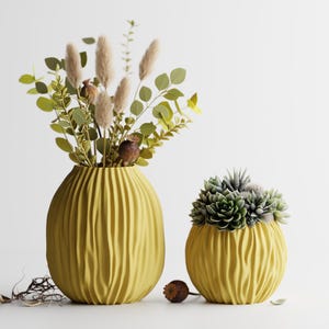 May include: Two yellow, textured vases. The larger vase holds a floral arrangement with green leaves and tan plumes. The smaller vase contains a succulent arrangement. Both vases have a ribbed design and sit on a white surface.