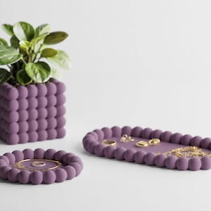 May include: A set of three purple matte home accessories featuring a bubble-textured design: a square planter containing a green plant, a small circular tray holding a gold ring, and a larger oval tray holding gold jewelry pieces.