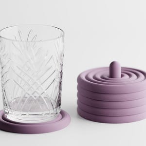 Könnte beinhalten: Ein klares Glas mit Muster steht auf einem lavendelfarbenen Untersetzer. Daneben ein Stapel lavendelfarbener Untersetzer mit einem zentralen Stift. Die Untersetzer bestehen aus weichem Material und schützen vor Wasserflecken.