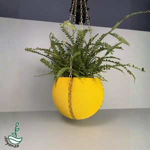 Op de afbeelding: Een hangende plantenbak met een heldergele, getextureerde, bolvormige pot. De pot wordt opgehangen door goudkleurige kettingen en is gevuld met groene varens. De plantenbak is ontworpen voor binnen of buiten.