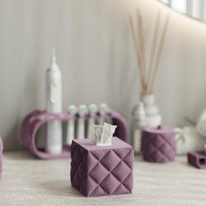 Puede incluir: Una funda cuadrada para caja de pañuelos con acabado mate en color malva y un patrón de diamantes acolchados. La funda tiene una abertura central en la parte superior por la que sobresalen pañuelos blancos. Combina con accesorios de baño.