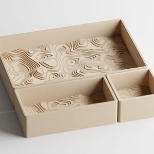 Può includere: Un set organizer da scrivania color beige composto da tre scomparti rettangolari di dimensioni variabili. Ogni scomparto presenta sulla base interna un motivo a rilievo stratificato che ricorda una mappa topografica.