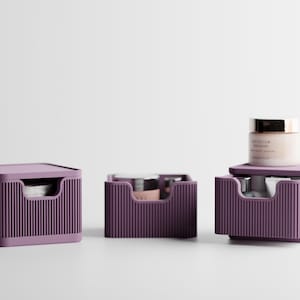Peut inclure: Trois organisateurs de rangement violets avec une texture nervurée verticale sur les façades des tiroirs. Un organisateur est fermé, un est ouvert avec un petit pot à l'intérieur, et un est ouvert avec un pot posé dessus. Poignées découpées.