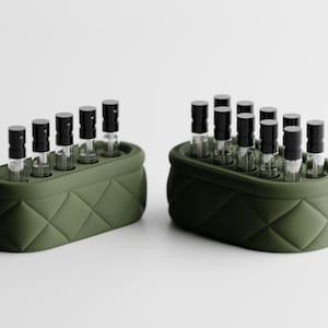 May include: Two olive green perfume sample holders, each holding multiple small glass vials with black caps. The holders have a quilted design. The vials are arranged in an organized manner, suggesting a fragrance collection or display.