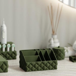 May include: A set of dark olive green bathroom accessories featuring a quilted diamond pattern. The collection includes a multi-compartment organiser, a toothbrush holder, a tissue box cover, a small lidded canister, and a rectangular soap dish.