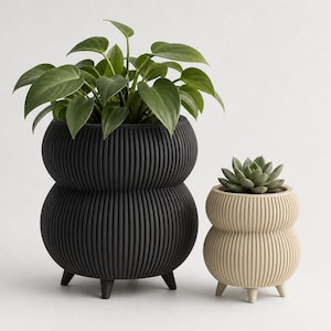 May include: A large black ribbed planter with a rounded double-tier shape and three small feet, containing a green leafy plant, next to a smaller beige ribbed planter with a matching shape and three feet, containing a green succulent.