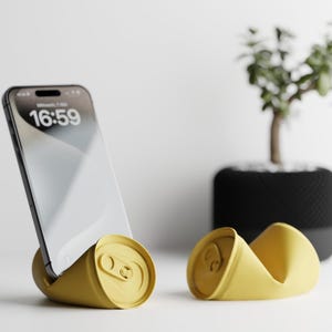 Könnte beinhalten: Ein gelber Telefonständer aus einer zerdrückten Dose, der ein Smartphone mit der Uhrzeit 16:59 hält. Daneben befindet sich ein weiterer gelber Dosenförmiger Ständer. Eine kleine Topfpflanze ist im Hintergrund.