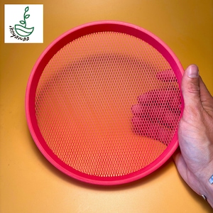 May include: A bright pink sieve with a diamond mesh pattern. The sieve has a circular shape and is held by a hand. The background is a solid yellow color. The sieve is likely used for straining or sifting ingredients in the kitchen.