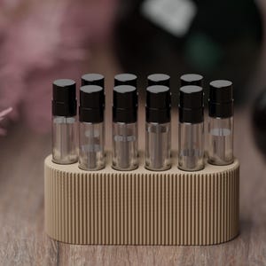 Könnte beinhalten: Ein Set aus sechs klaren Glasfläschchen mit schwarzen Kappen, angeordnet in zwei Reihen zu je drei Stück, gehalten in einem beigen, ovalen Ständer mit vertikaler Rippenstruktur. Die Fläschchen sind transparent und leer gestaltet.