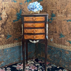 May include: A dark wood antique cabinet with four drawers, featuring ornate inlaid designs in shades of brown and gold. The cabinet has delicate legs and decorative metal accents. A bouquet of blue flowers sits atop the cabinet.