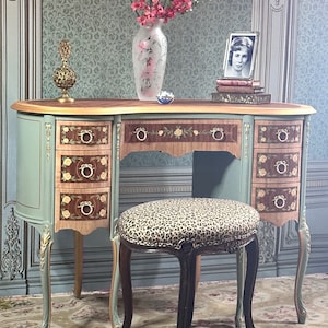 May include: An antique vanity desk with a matching stool. The desk is painted in a soft blue-green with gold accents and floral inlay. A leopard print stool sits in front. A vase of pink flowers and a framed photo are on the desk.