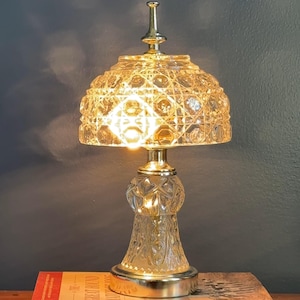 May include: A vintage table lamp with a clear, faceted glass base and shade. The lamp has a gold-colored metal stem and finial. It sits on top of a stack of books with titles including "The Great Hound Match of 1905" and "Centered Riding".