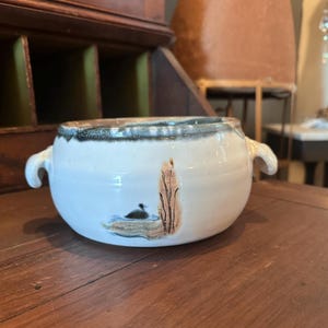 May include: A white ceramic bowl with two handles and a painted scene of a duck on water with reeds. The rim is dark blue and brown. The bowl is sitting on a wooden surface.
