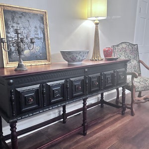 May include: A dark wood console table with ornate carvings and a black painted finish. A gold-framed painting, a floral bowl, and a gold lamp with a cream shade sit on top. A vintage chair with floral upholstery is to the right.