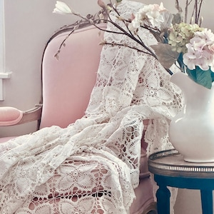 May include: A pink upholstered chair draped with a white lace textile. A white pitcher with flowers sits on a small blue side table. The scene is soft and romantic, with a vintage aesthetic.