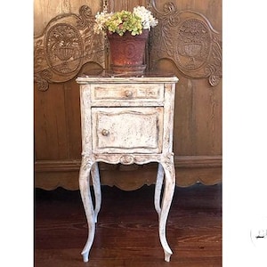 May include: A distressed white bedside table with a drawer and cabinet door, featuring ornate curved legs. A terracotta pot with flowers sits on top. The table is approximately 28 inches tall.