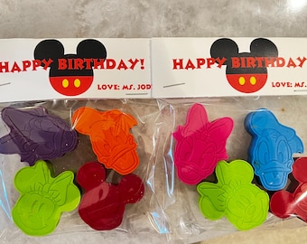 May include: Two clear plastic bags with Mickey Mouse themed birthday tags. Each bag contains five colorful wax crayons shaped like Mickey Mouse and Minnie Mouse characters. The tags read "Happy Birthday! Love: Ms. Jody".