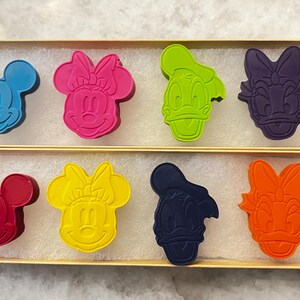 Op de afbeelding: Acht kleurrijke, figuurvormige kleurpotloden in een houten doos. De kleurpotloden zijn gevormd als Mickey Mouse, Minnie Mouse, Donald Duck en Daisy Duck. De kleurpotloden zijn gerangschikt in twee rijen van vier.